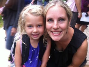  a sweet moment between Lucy and her mom, Joy, on Lucy’s first day of first grade. Lucy, with her curly blonde hair and a gentle smile, wears a purple dress with white stars and a checkered backpack. Joy, smiling brightly beside her, leans in close, her blonde hair pulled back. The background suggests an outdoor school setting, filled with excitement and new beginnings.